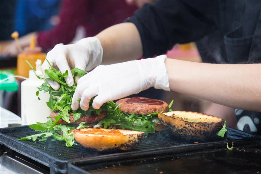 gloved hands adding garnish to a hamburger on a grill