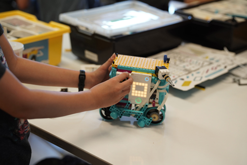 Youth Academy student adjusts part of a LEGO robot. The robot is a cube shape with wheels, and is on a white tabletop.