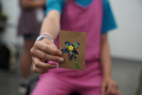 A child holding a piece of art work up to the camera. The art work is a small kraft paper card with a mixed media collage flower on it.