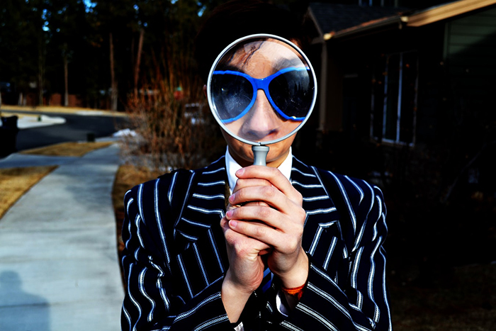 Person in striped blazer looks through magnifying glass in blue sunglasses